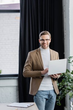 Bilgisayarlı gülümseyen işadamı panonun yanındaki kameraya bakıyor. 