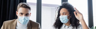 Tıbbi maske takmış Afro-Amerikan iş kadını meslektaşıyla konuşurken el kol hareketi yapıyor.