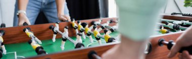 partial view of interracial business colleagues playing table football, blurred foreground, banner clipart