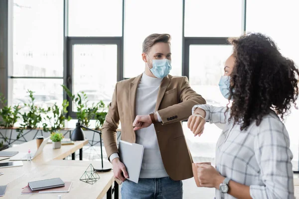 interracial business partners in medical masks doing elbow greeting in office