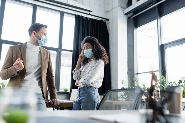 african american businesswoman in medical mask holding hand near head during conversation with colleague