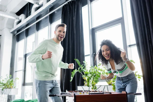 excited interracial business partners showing win gesture near table football