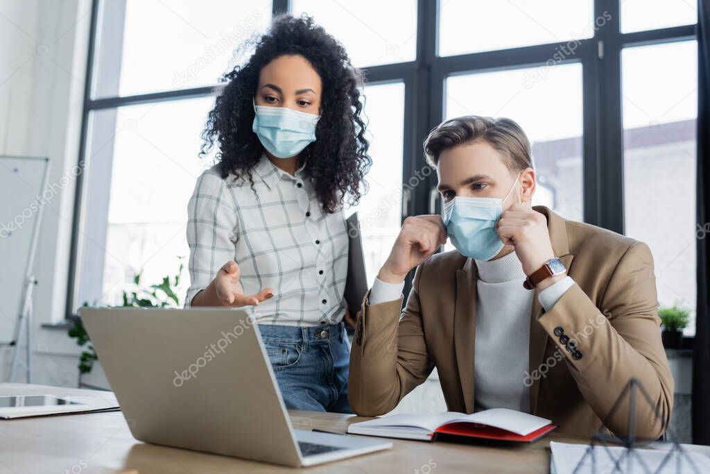 african american businesswoman in medical mask pointing at laptop near business partner