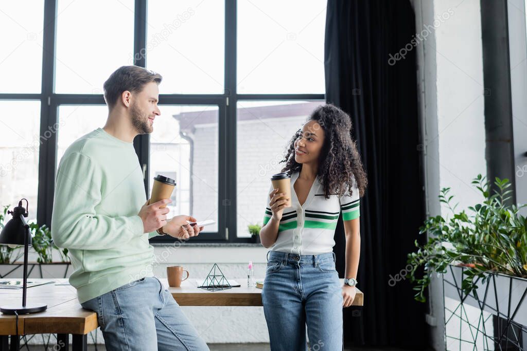 happy interracial business partners standing with coffee to go during conversation in office