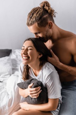 Yatakta yastık tutan neşeli kız arkadaşının kafasını öpen seksi adam. 