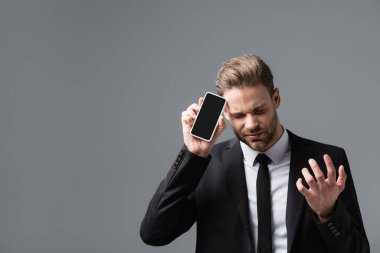frustrated manager gesturing while showing cellphone with blank screen isolated on grey clipart