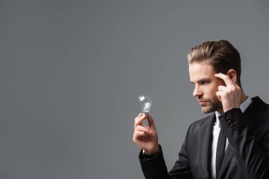 pensive businessman touching head while looking at light bulb isolated on grey clipart