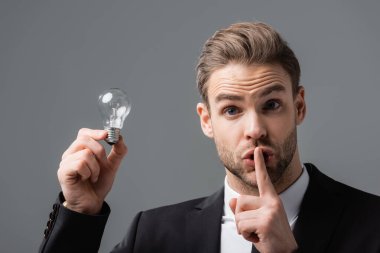 thrilled businessman holding light bulb while showing secret gesture isolated on grey clipart