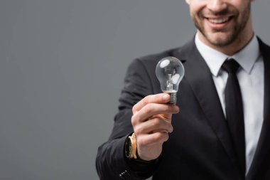 cropped view of smiling businessman showing light bulb on blurred background isolated on grey clipart