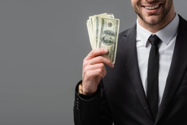 partial view of smiling businessman in black suit holding money isolated on grey clipart
