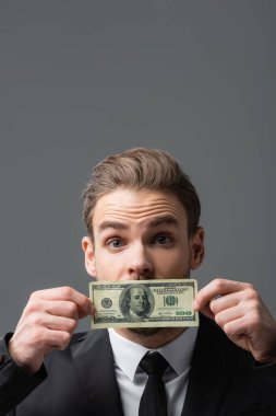 Gri üzerine izole edilmiş dolar banknotuyla ağzını kapayan heyecanlı iş adamı