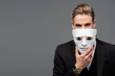 Gri renkli kameraya bakarken yüzünün yanında maske tutan iş adamı