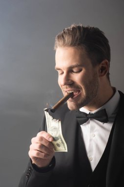 wealthy businessman lighting cigar with hundred dollar banknote on grey background with smoke clipart
