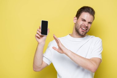 man feeling disgust and showing refuse gesture while holding cellphone with blank screen on yellow