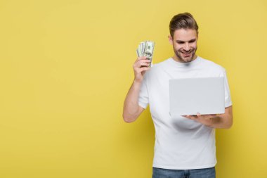 pleased man holding dollar banknotes while looking at laptop on yellow