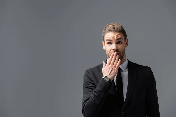 confused businessman covering mouth with hand while looking at camera isolated on grey