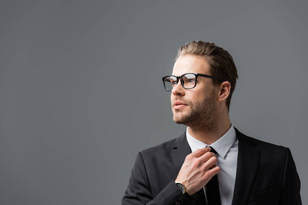 confident businessman in eyeglasses adjusting tie while looking away isolated on grey