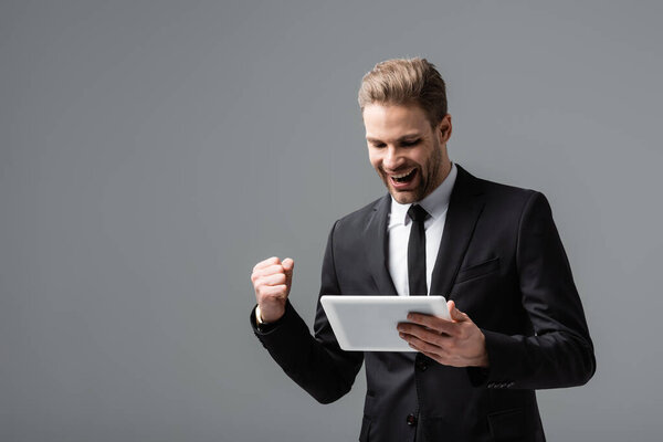 взволнованный бизнесмен показывает победу жест в то время как проведение цифровой планшет изолирован на серый