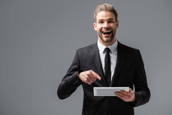 excited manager pointing at digital tablet while looking at camera isolated on grey