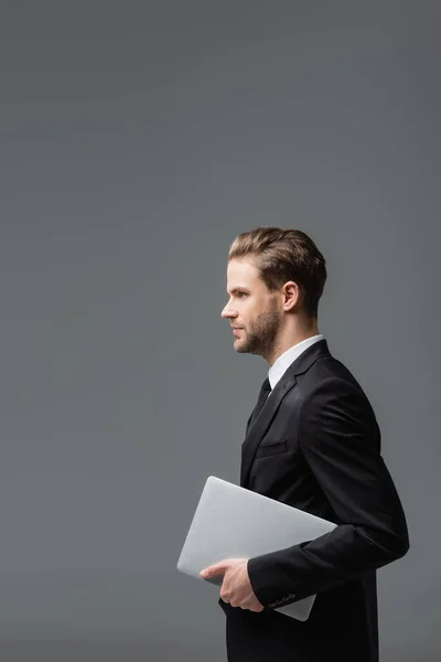 side view of young executive manager holding laptop isolated on grey