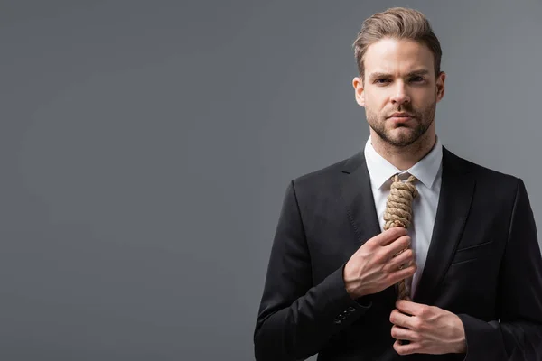 frustrated businessman looking at camera and touching tie made of rope isolated on grey