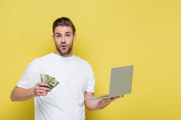 astonished man with laptop and dollar banknotes looking at camera on yellow