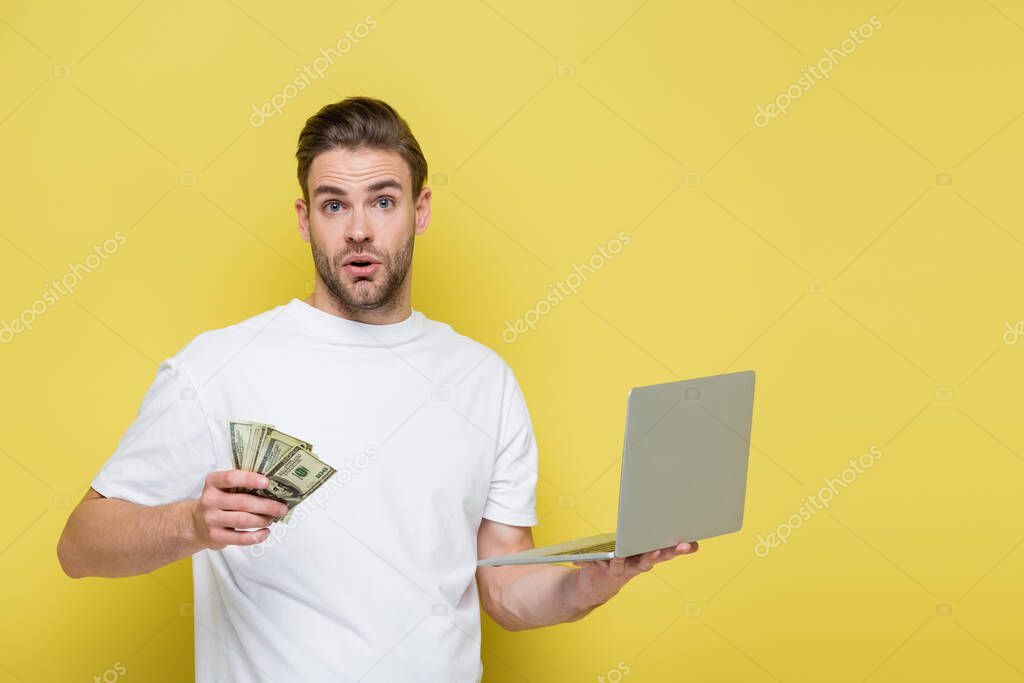 astonished man with laptop and dollar banknotes looking at camera on yellow