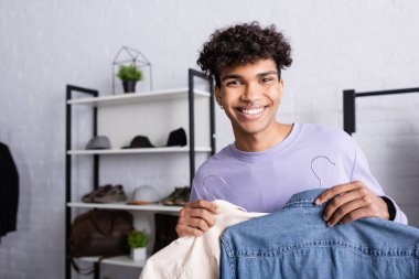 Arfican Amerikalı iş adamı askılarda gömlek tutuyor ve sergi salonunda kameraya gülümsüyor. 