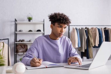 Young african american businessman using laptop and writing on notebook in showroom with clothes  clipart