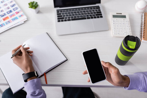 Обрезанный вид владельца африканского американского шоурума, держащего смартфон с чистым экраном и пишущего на ноутбуке 