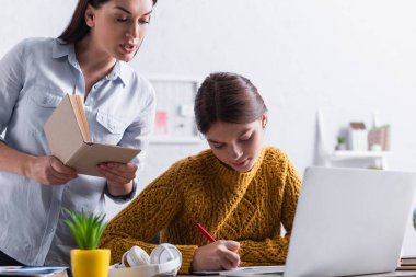 Genç kız ödevlerini yaparken annesinin yanında kitap ve dizüstü bilgisayarla yazıyor.