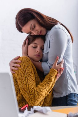 Endişeli anne, dizüstü bilgisayarın yanında gözleri kapalı, üzgün genç kızı kucaklıyor.