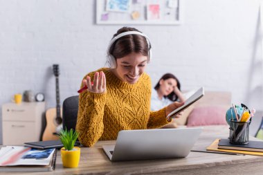 Telsiz kulaklıklı mutlu genç kız dizüstü bilgisayarın yanında elinde defter ve kalem tutuyor.