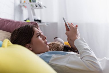Akıllı telefon kullanan ve oyuncak ayı tutan üzgün bir genç kız.