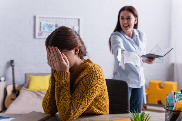 teenage girl covering face with hands near angry mother tearing papers in notebook