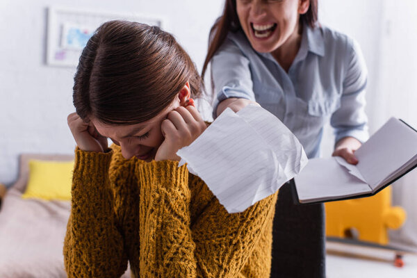 scared teenage girl near angry mother tearing papers in notebook