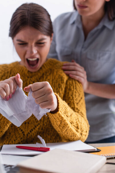 angry teenage girl tearing paper and screaming near worried mother