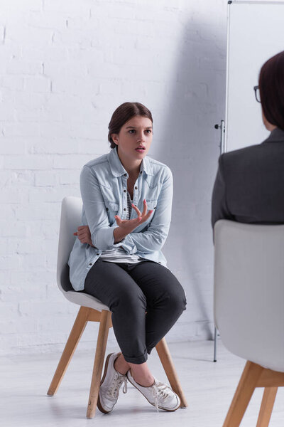 worried teenage girl sitting and gesturing while looking at psychologist on blurred foreground 