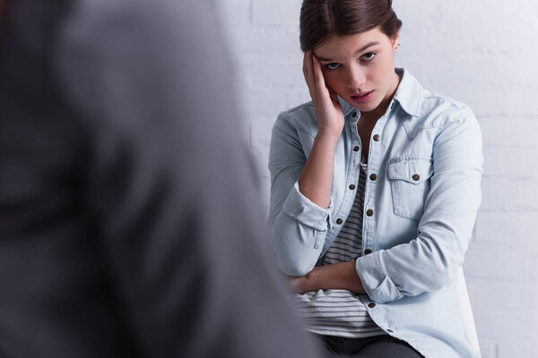 worried teenage girl looking at camera near psychologist on blurred foreground 