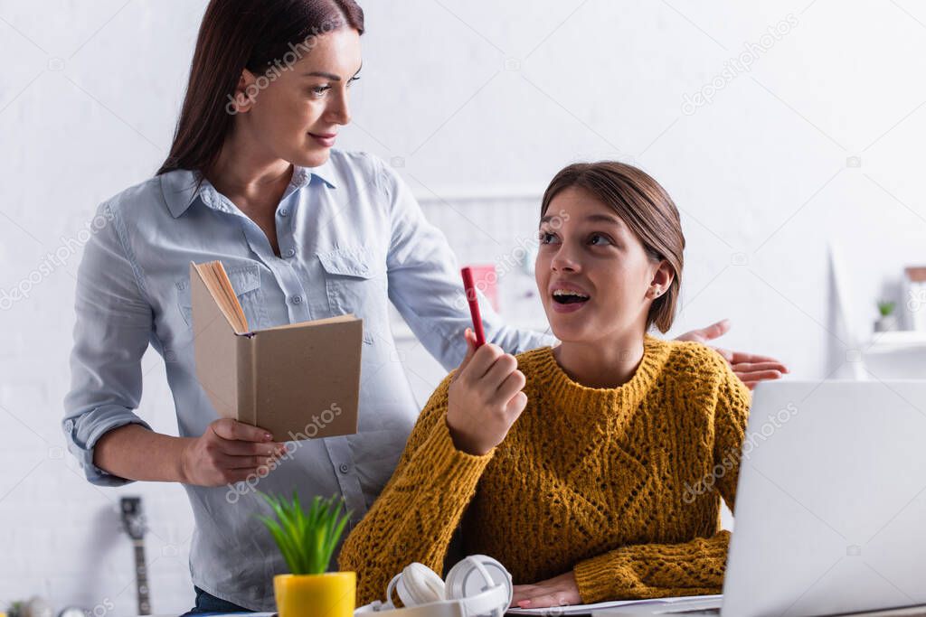 teenage girl thinking and holding pen while doing homework near mother with book 