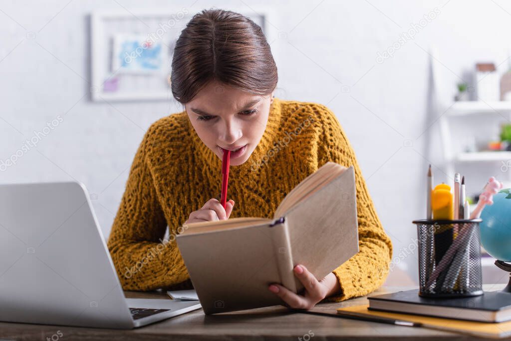 thoughtful teenage girl reading book and holding pen while studying online