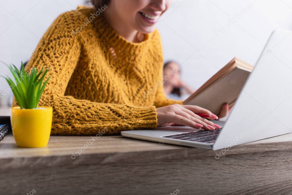 cropped view of cheerful teenage girl typing on laptop keyboard and holding book