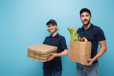 Pizza kutularıyla neşeli çok kültürlü kuryeler ve mavi üzerine izole edilmiş yiyeceklerle dolu kağıt torbalar.