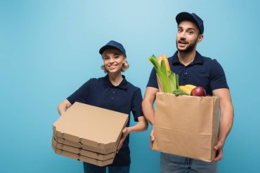 Pizza kutularıyla neşeli ırklar arası kuryeler ve mavi üzerine izole edilmiş yiyecek dolu kağıt torbalar.