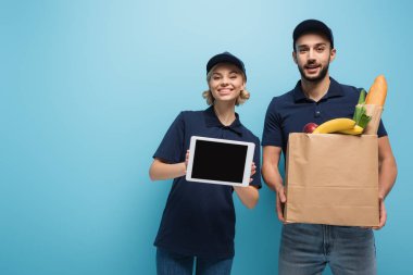 Neşe dolu ırklar arası kuryeler ellerinde boş ekranlı dijital tablet ve mavi üzerine izole edilmiş yiyecek dolu kağıt torba tutuyorlar.