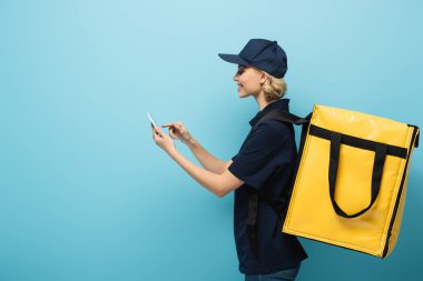 Gülümseyen kuryenin yan görünüşü, termo torba ile mavi cep telefonunu gösteriyor.