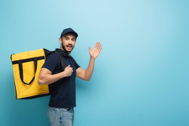 Termo çantalı mutlu Müslüman teslimatçı mavi kameraya el sallıyor.