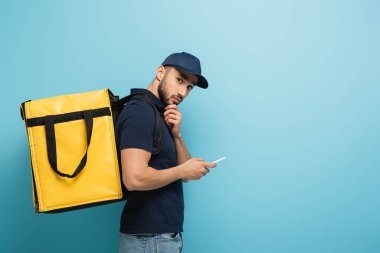 Termo sırt çantalı düşünceli Arap kurye mavi üzerinde moblie telefon tutuyor
