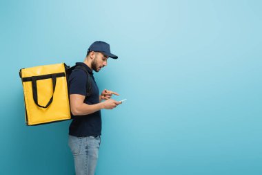 Mavi renkteki akıllı telefonu gösteren termo sırt çantalı Arap kuryenin yan görüntüsü