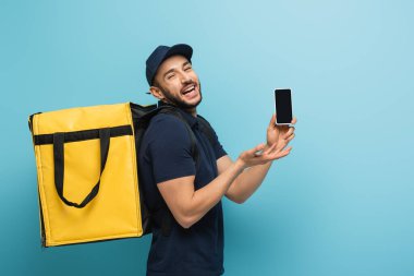 Heyecanlı Müslüman kurye, mavi üzerine akıllı telefonu işaret ederken gülüyor.